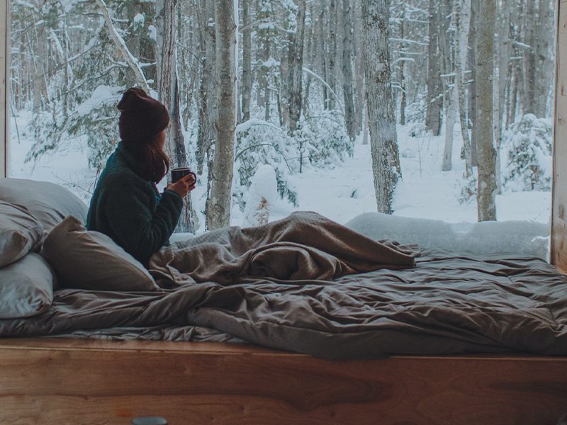 Kvinde sidder med kaffe under tæppe og kigger ud på sneklædt skov